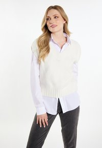 Gilet bianco lavorato a maglia con scollatura a V, tessuto texturizzato e orlo a coste, indossato sopra una camicia bianca con colletto e abbinato a jeans grigi scuri.