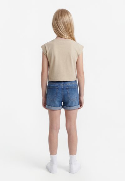 Girl with long blonde hair wearing beige sleeveless top, blue rolled-up denim shorts, white socks, and white sneakers, standing facing away.