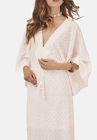 Vestido blanco con estampado, escote en V profundo, mangas amplias y cintura con cordón. Presenta bordados circulares y una tela ligera y texturizada.