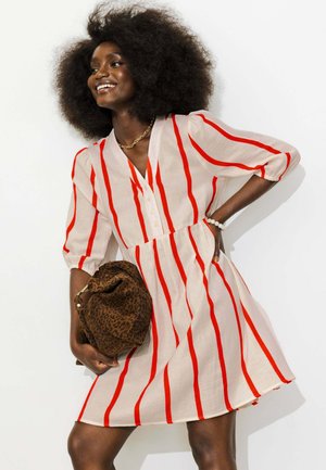Femme avec un grand afro portant une robe beige à rayures rouges verticales, tenant un sac marron à imprimé léopard, souriante et posant devant un fond blanc.