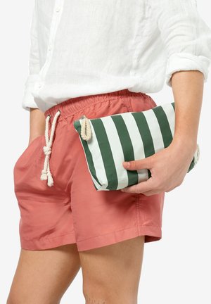 Clutch a righe in tessuto verde e bianco, con una texture morbida e dettagli in corda. Tenuto in mano accanto a pantaloni corti rosa e una camicia bianca.