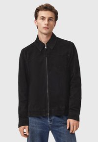 Mand iført en sort ruskinds zip-jakke med krave og blå jeans, der står ansigtet fremad mod en enkel lys baggrund.