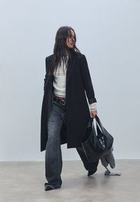 Pitkä musta takki, jossa on lovetut kaulukset, yhdistetty valkoisiin raidallisiin neuleeseen, tummiin farkkuihin ja mustaan nahkakassiin. Yksinkertainen muotoilu ja tekstuuri.