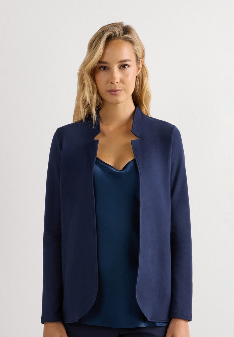 Mujer rubia con una chaqueta azul marino abierta sobre un camisón de satén azul oscuro, en pie sobre un fondo blanco liso.