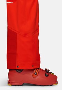 Pantalons de ski rouges avec un panneau d'accent texturé, associés à des bottes de ski rouges et noires avec des éléments orange et une semelle en caoutchouc.
