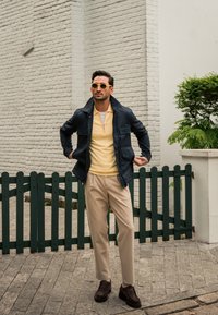 Chaqueta navy sobre un polo amarillo claro, pantalones beige y zapatos marrón oscuro. Gafas de sol redondas amarillas completan el look. Pared de ladrillo y cerca verde de fondo.