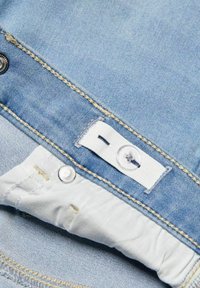 Svetlemodrý džínovinový materiál s bílou nášivkou, žlutým šitím a stříbrným detailem knoflíku poblíž pasu. Jemná textura a jednoduchý design.