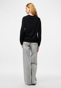 Moteris su ilgais šviesiais plaukais, vilkinti juodą ilgomis rankovėmis megztinį ir šviesiai pilkas plačias kelnes, stovi ir atsisukusi į šalį ant balto fono.