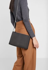 Czarna torebka kopertówka z prążkowanej tkaniny z srebrnym łańcuszkiem. Teksturowany materiał, prostokątny kształt i zamek błyskawiczny. Stylizowana z brązowymi spodniami z wysokim stanem.