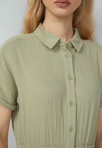 Camicia verde chiaro, con bottoni e realizzata in tessuto testurizzato. Presenta un colletto e maniche corte, con delicate pieghe e un taglio rilassato.