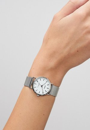 Håndled med et ur med sølvfarvet mesh-rem, hvid urskive, sorte timemarkeringer, slanke sorte visere og en lille sekundviser ved klokken 6.