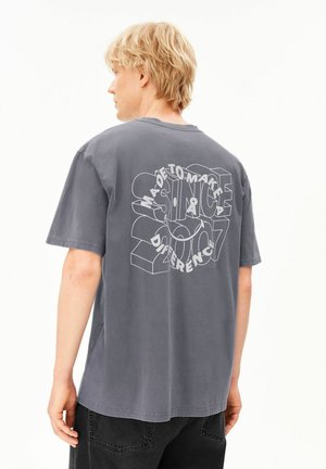 Blonder Mann trägt ein lockeres graues T-Shirt mit weißem 3D-Text auf dem Rücken mit der Aufschrift "Made to Make a Difference" und schwarze Hosen.
