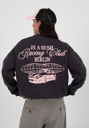 Sweat-shirt noir avec un graphisme rose représentant une voiture et un globe, accompagné du texte "IN A RUSH Racing Club BERLIN" affiché de manière bien visible.