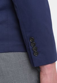 Mörkblå kostyms ärm med fyra svarta knappar längs manschetten, i kombination med grå strukturerade byxor och synlig hud på handleden.