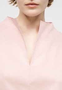 Κοντινό πλάνο ενός ατόμου που φοράει ανοιχτό ροζ τοπ με λαιμόκοψη σε σχήμα V και χρυσούς κρίκους σκουλαρίκια.