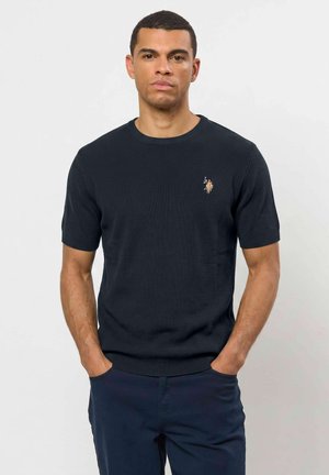 Homme portant un polo à manches courtes bleu marine en maille avec un petit logo brodé sur la poitrine, debout, mains dans les poches, devant un fond uni.