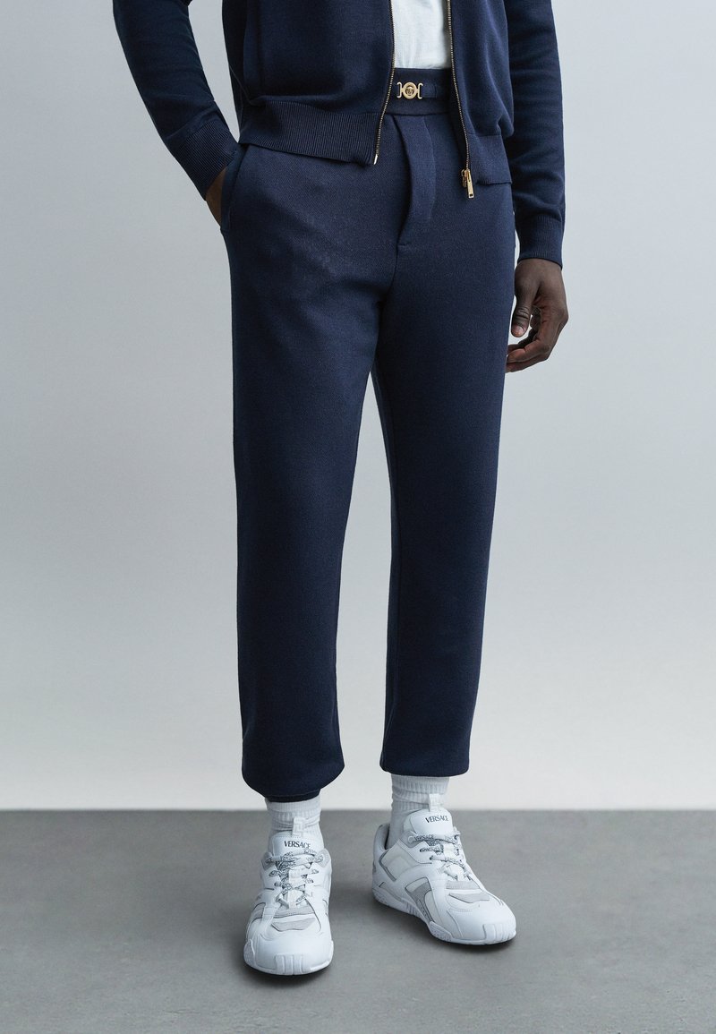 Marinblå stickade träningsbyxor med zipficka, kombinerade med vita sneakers med tydlig logotyp och texturerad design.