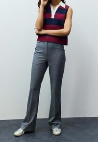 Femme portant un pantalon gris, des baskets blanches et un haut sans manches en maille rayé bordeaux et bleu marine, debout contre un mur uni.