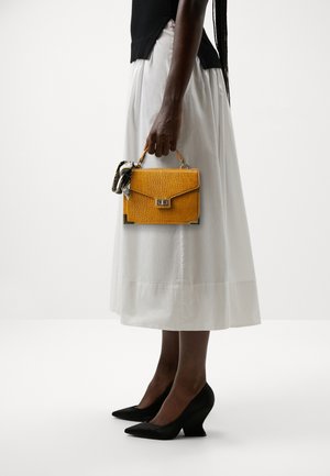 Personne en jupe blanche et haut noir tenant un petit sac à main jaune texturé avec un foulard, portant des escarpins noirs pointus sur un fond blanc.