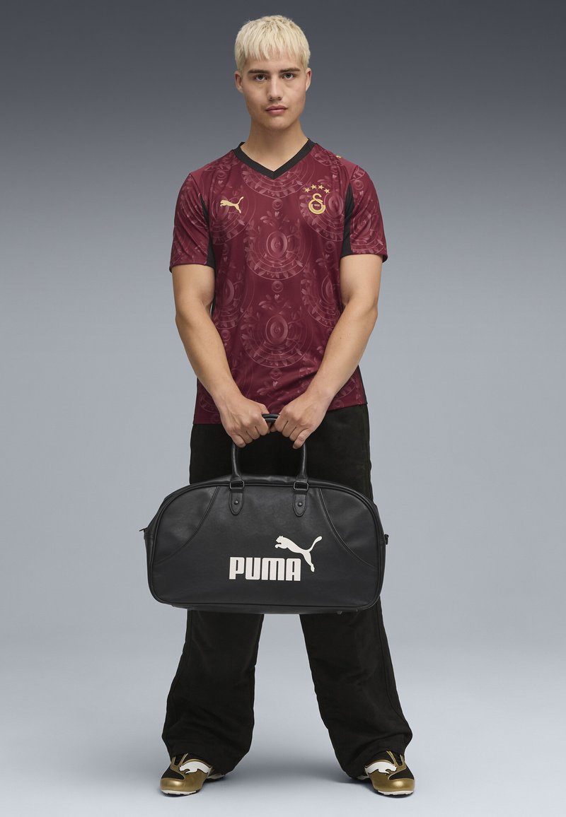 Bordeauxfarbener Trikot mit kreisförmigem Muster, schwarzen Ärmeln und goldenen Akzenten. Schwarze Lederhandtasche mit weißem "PUMA"-Logo, von einer Person gehalten.