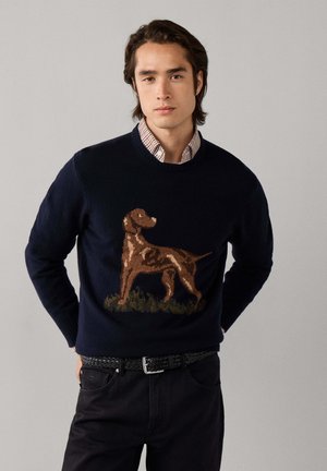 Modelo masculino que lleva un jersey azul marino con diseño de perro, camisa de cuello a cuadros, pantalones negros, cinturón negro y botas de senderismo marrones, posando frente a un fondo gris.