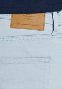 Světle modrá denimová tkanina s koženou nášivkou v hnědé barvě, na které je vyražený text. Další šití podrobností podél kapsy.