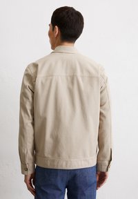 Beige cotton jacket with a simple design, featuring a straight cut, back yoke, and button accents at the cuff. Worn over blue denim pants.