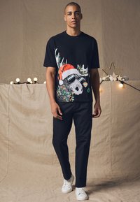 Black t-shirt featuring a colorful graphic of a skull with antlers, wearing a red Santa hat and sunglasses. Paired with dark pants and white shoes.