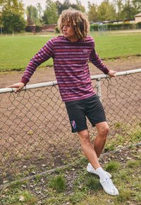 Burgundi és tengerészkék csíkos hosszú ujjú póló, fekete rövidnadrág logóval, fehér sportcipő, füves mező és láncos kerítés előtt.