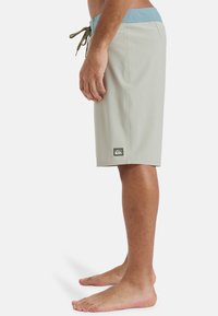 Svetlo šedé plážové šortky s modrým pásom, sťahovacou šnúrkou a nášivkou loga na okraji; hladký materiál a dizajn po kolená.