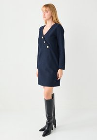 Robe bleu marine avec col en V, manches longues et trois boutons dorés. Confectionnée en tissu texturé, associée à des bottines noires montantes à talons.