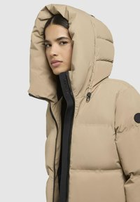 Giacca a vento beige con un ampio cappuccio imbottito, zip nera e dettagli con logo. Presenta una texture trapuntata e un colletto alto per un maggior calore.
