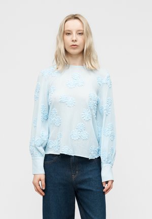 Blondínka v svetlomodrej blúzke s textúrovanými kvetinovými vzormi a tmavomodrých vysokých džínsach stojaca pred jednofarebným pozadím.