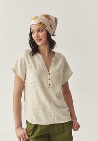 Beige geribbelde blouse met bloemenborduursel, een V-hals met drie knopen, korte mouwen, gecombineerd met groene broeken en een bloemen sjaal.