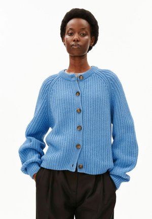 Person trägt eine blaue grob gestrickte Strickjacke mit braunen Knöpfen und schwarze Hose, steht mit Händen in den Taschen vor einem weißen Hintergrund.