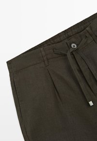 Mørke olivengrønne bukser med elastisk talje, knaplukning, bæltestropper og justerbar stofsnor på hvid baggrund.