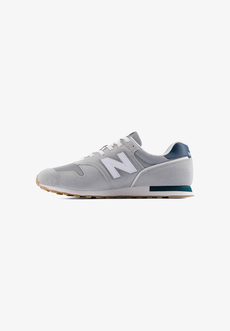 Zapatilla New Balance gris y blanca con parte superior de ante y malla, cierre de cordones y suela de goma color caramelo, mostrada de perfil sobre fondo blanco.