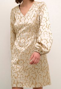 Beige klänning med V-ringning, som har ett guld abstrakt mönster, långa puffärmar med elastiska manschetter och en figurnära silhuett.