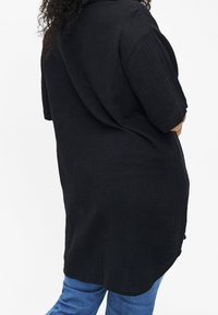 Chemise noire oversize en tissu texturé et côtelé. Caractéristiques des manches courtes et un ourlet courbé. Associée à un jean bleu.