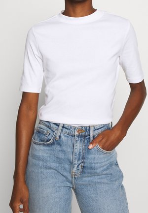 Persoon die een effen wit T-shirt met korte mouwen draagt, ingestopt in een high-waisted lichtblauwe spijkerbroek, met één hand in de zak.