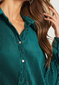 Blusa teal a coste con collo a camicia, bottoni dorati e maniche lunghe. Texture morbida con una leggera lucentezza. Inquadratura ravvicinata del tessuto.