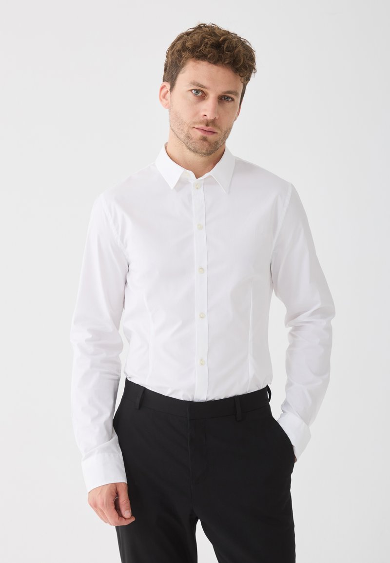 Moški z kodrastimi lasmi, oblečen v belo srajco s kragnico in črne hlače, stoji ob enostavni svetli ozadju z eno roko v žepu.