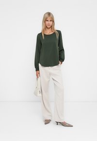 Mujer vestida con una blusa de manga larga verde oscura, pantalones de pierna ancha color crema, tacones topo y sosteniendo un bolso blanco contra un fondo blanco.