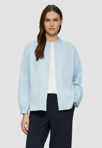 Lichtblauw button-up shirt met een ronde hals, lange mouwen en een ontspannen pasvorm. Gemaakt van zachte stof met een subtiele textuur. Gekleed met donkere broeken.