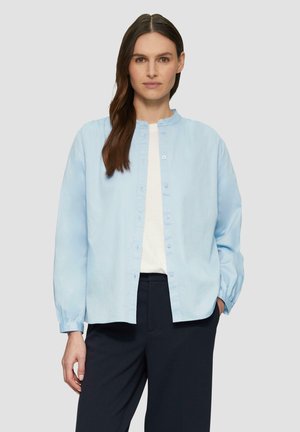 Lichtblauw button-up shirt met een ronde hals, lange mouwen en een ontspannen pasvorm. Gemaakt van zachte stof met een subtiele textuur. Gekleed met donkere broeken.