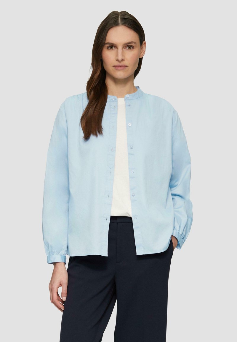 Lichtblauw button-up shirt met een ronde hals, lange mouwen en een ontspannen pasvorm. Gemaakt van zachte stof met een subtiele textuur. Gekleed met donkere broeken.