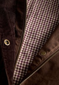 Close-up of a brown jacket showing a metal zipper, snap button, corduroy fabric, and a purple and white checkered inner lining.