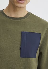 Oliwkowy polarowy sweatshirt z granatową kieszenią na lewej piersi. Posiada okrągły dekolt i miękką teksturę.