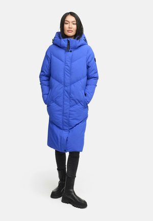 Manteau long bleu en duvet avec une capuche, présentant un design matelassé en chevrons, une fermeture éclair à l'avant et des poches latérales. Associé à un pantalon noir et des bottes.