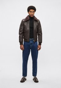 Brown leather bomber jacket with a fur collar, worn over a black turtleneck, paired with blue denim jeans and dark brown loafers.